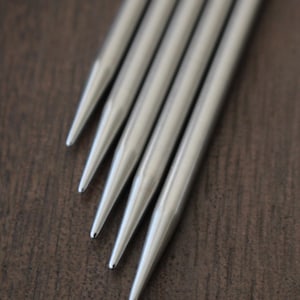 以下が含まれることがあります： 茶色の木製の表面に、先のとがった先端を持つ銀色の編み針が4本、扇形に配置されています