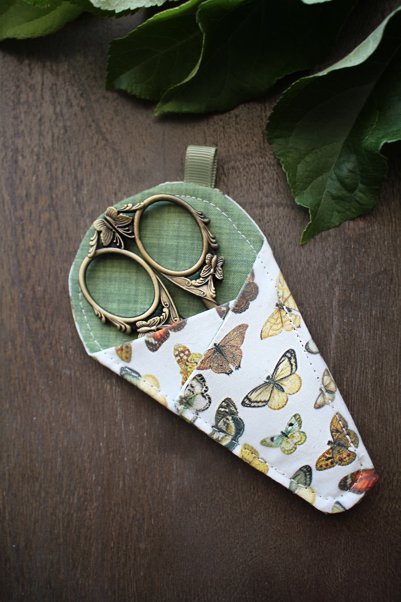 May include: A green fabric scissor case with a butterfly print and a pair of antique-style scissors with ornate handles.