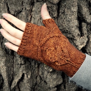 May include: A pair of brown fingerless gloves with a leaf pattern knitted into the fabric. The gloves are worn on a hand resting against a tree trunk.