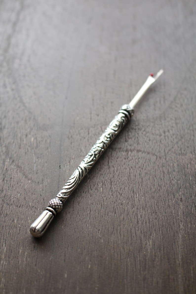 May include: A silver thread cutter with an ornate, detailed handle. The handle is decorated with a floral pattern.