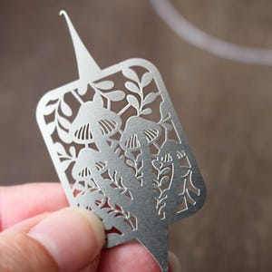 May include: A silver-colored metal tool with a rectangular cutout design featuring mushrooms and leafy stems. The tool has a pointed top and a small hook at the bottom, with a flat, brushed finish and intricate laser-cut details throughout the surface.
