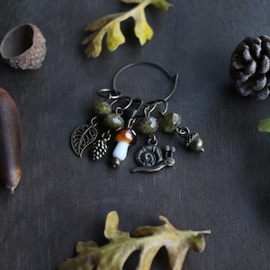 May include: A brown metal stitch marker with a variety of charms, including a mushroom, a snail, a leaf, and acorns. The charms are attached to a circular metal ring.