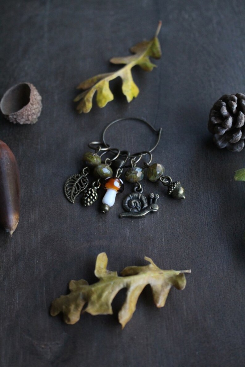 May include: A brown metal stitch marker with a variety of charms, including a mushroom, a snail, a leaf, and acorns. The charms are attached to a circular metal ring.