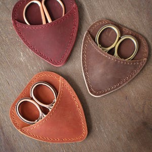 May include: Three leather scissor cases in shades of red, brown, and tan, each holding a pair of small scissors. The cases are heart-shaped with stitched edges, and the scissors have gold or silver-colored handles.