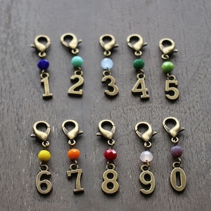 Könnte beinhalten: Zehn bronzene Zahlenanhänger mit bunten Perlen. Die Zahlen sind 1 bis 10. Die Perlen sind blau, türkis, hellblau, grün, grün, gelb, orange, rot, rosa und lila.