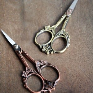 May include: Two pairs of decorative scissors with ornate butterfly designs. One pair is gold with a silver blade, and the other pair is copper with a silver blade.