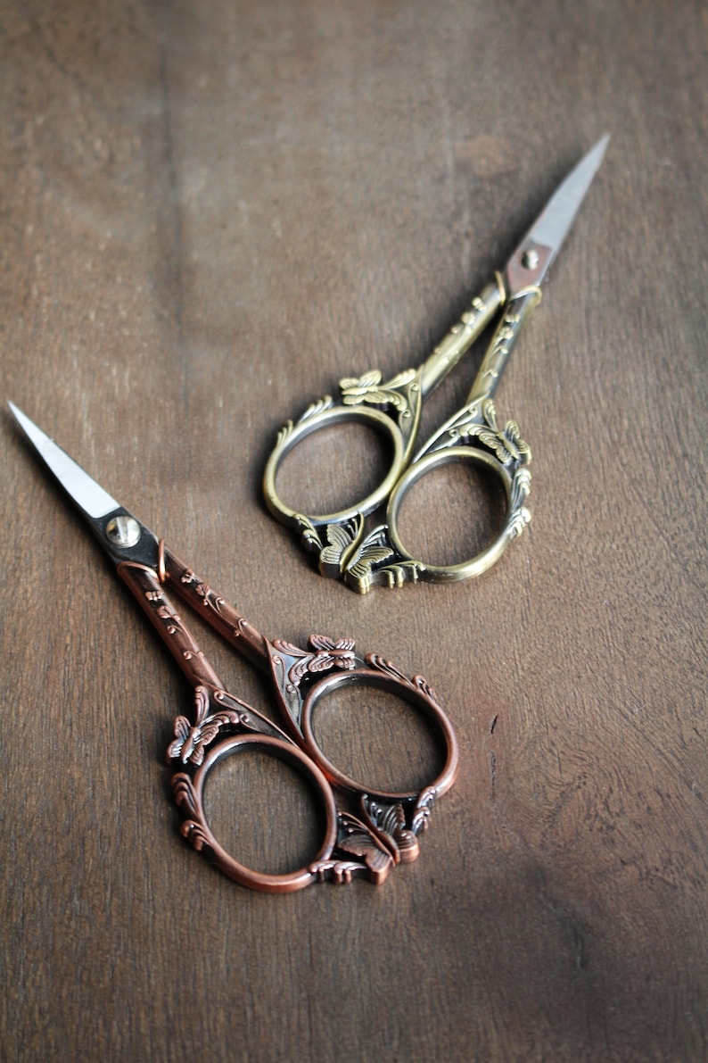 May include: Two pairs of decorative scissors with ornate butterfly designs. One pair is gold with a silver blade, and the other pair is copper with a silver blade.