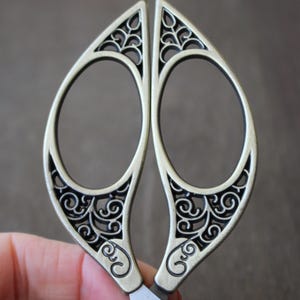 May include: Silver-toned decorative scissors with an intricate, filigree design. The handles are shaped like leaves and have a delicate, openwork pattern.