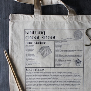May include: A beige tote bag with a black and white knitting cheat sheet printed on it. The cheat sheet includes knitting abbreviations and techniques. The bag has two wooden knitting needles.