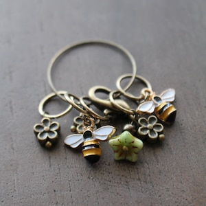 May include: A set of four gold-toned stitch markers with bee and flower charms. The markers are connected by a ring.