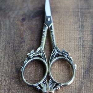 May include: Antique silver-toned scissors with ornate butterfly and floral designs. The scissors have a pointed tip and are closed.