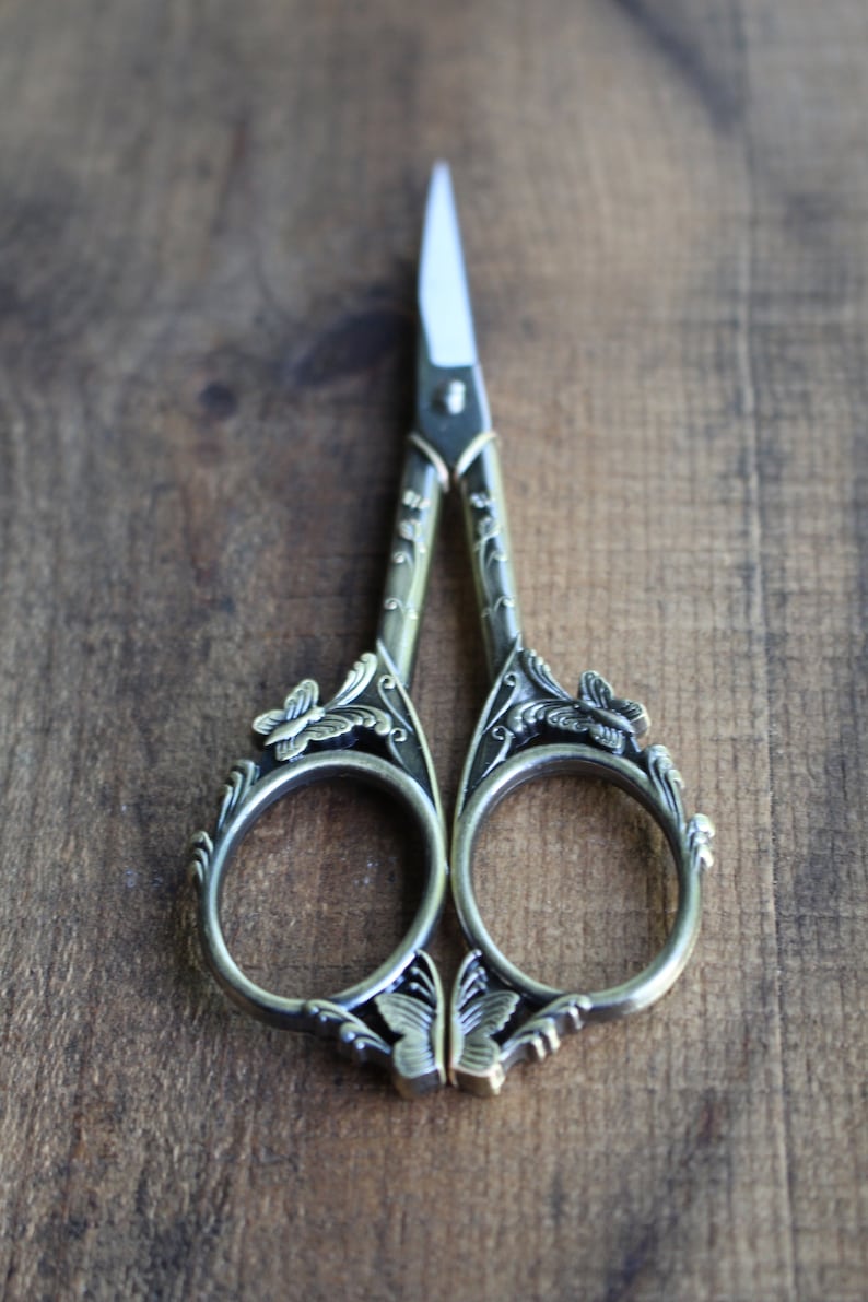 May include: Antique silver-toned scissors with ornate butterfly and floral designs. The scissors have a pointed tip and are closed.