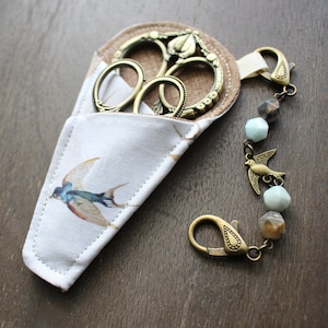 Puede incluir: Un estuche para tijeras de tela blanca con un diseño de golondrina y unas tijeras doradas con un diseño floral. El estuche tiene una solapa de tela marrón y una cadena dorada con un dije de pájaro y cuentas.