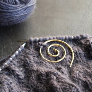 May include: A close-up of a gray knitted fabric with a gold spiral stitch marker. The fabric is soft and fuzzy, and the stitch marker is a simple, elegant design.