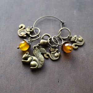 May include: A gold-toned metal knitting stitch marker with a ring and multiple charms, including squirrels and acorns. The charms are attached to the ring with jump rings.