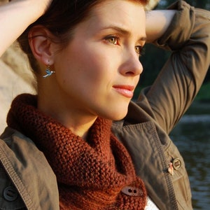 May include: A woman with brown hair is wearing a brown knitted cowl neck scarf with wooden buttons. She is also wearing a brown jacket and a white shirt.