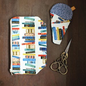 May include: Two white fabric pouches with a book print design. One pouch is a small, triangular shape and the other is a larger, rectangular shape. Both pouches have zippers. A pair of gold-colored scissors is also in the image.
