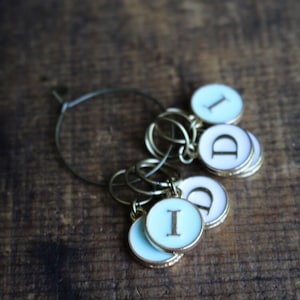 May include: A set of six silver and gold stitch markers with blue and white enamel. The markers are in the shape of circles with the letters "I" and "D" on them.