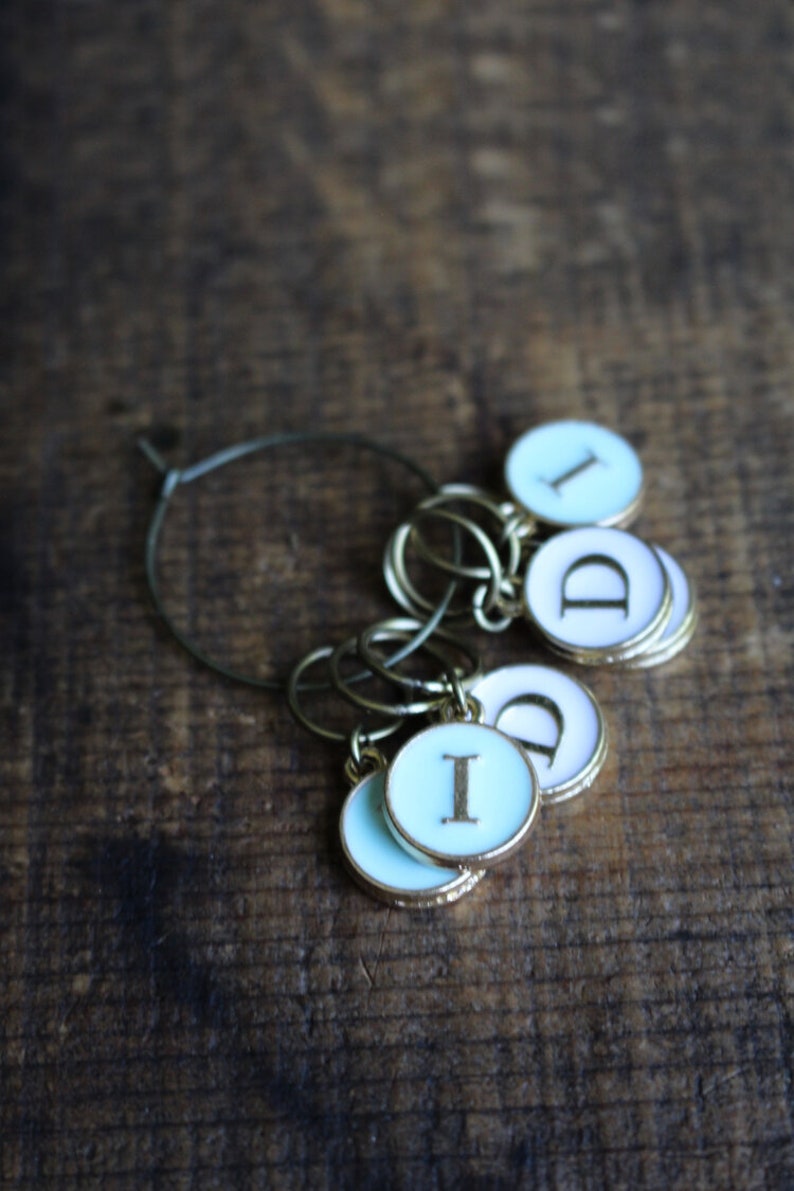May include: A set of six silver and gold stitch markers with blue and white enamel. The markers are in the shape of circles with the letters "I" and "D" on them.