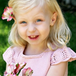 Könnte beinhalten: Ein junges Mädchen mit blonden Haaren lächelt, während es einen Strauß rosa Blumen hält. Sie trägt ein rosa Strickkleid mit einem zarten Spitzenmuster.