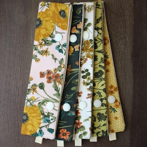 May include: Five rectangular fabric straps featuring various floral patterns in shades of yellow, green, white, and black. Each strap includes two white plastic snap buttons and a small beige fabric loop at the bottom edge for hanging or attachment.