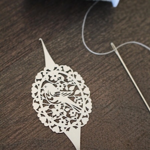 May include: An off-white die-cut paper ornament featuring an oval frame with a bird and floral motif, pointed ends, and a small hook. A spool of white thread and a silver sewing needle are positioned nearby for scale and context.