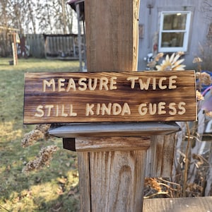 May include: A rectangular wooden sign with the text "MEASURE TWICE STILL KINDA GUESS" carved into the surface. The sign is mounted on a wooden post, with a natural wood grain and a dark brown finish.