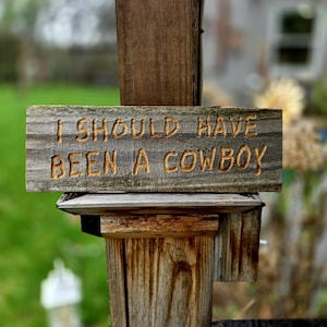 May include: A weathered wooden sign with the carved text "I SHOULD HAVE BEEN A COWBOY" is mounted on a wooden post. The sign is rectangular and has a rustic, aged appearance. The text is in a simple, sans-serif font.