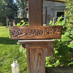 以下が含まれることがあります： 「MR SANDERZ」の文字が彫られた長方形の木製看板。看板は濃い茶色の木材でできており、木製の柱に取り付けられています。背景には緑の草や葉があります。