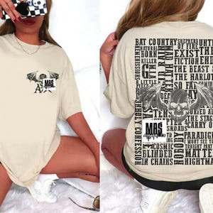 Peut inclure: T-shirt beige à manches courtes avec un motif graphique à l'avant et à l'arrière. Le motif avant présente un design de chauve-souris et de crâne avec les lettres "MAS". Le dos a un design de crâne et d'ailes avec du texte.