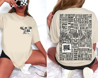 May include: Beige short-sleeved t-shirt with a graphic on the front and back. The front graphic features a bat and skull design with the letters "MAS". The back has a skull and wing design with text.