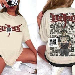 May include: Cream-colored t-shirt featuring the text "Kid Rock" in a maroon, stylized font. The back of the shirt displays a collage of text and an image of Kid Rock. The shirt has short sleeves and a relaxed fit.