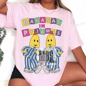 May include: Pink t-shirt with a graphic of two cartoon bananas wearing blue and white striped pajamas. The text "Bananas in Pajamas" is written in colorful block letters above the bananas. The text "Mas Apparel Co" is written in white letters on a black background below the bananas.