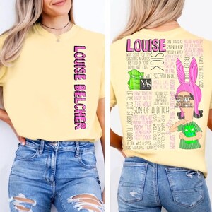May include: A pale yellow t-shirt featuring the text "LOUISE BELCHER" in pink and black on the front. The back displays a graphic of a character with pink bunny ears, a green shirt, and extensive black text quotes from the show Bob's Burgers.