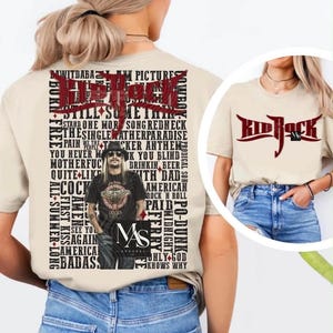 May include: A beige short-sleeve t-shirt featuring a large graphic on the back with the text Kid Rock in red, a central image of the artist, and a list of song titles in black. The front of the shirt displays the red Kid Rock logo on the chest.
