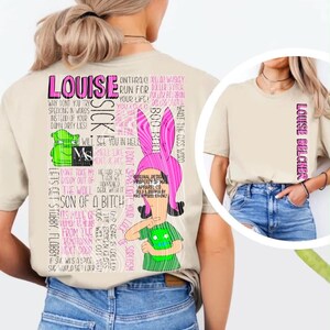 May include: A beige t-shirt featuring a large graphic on the back with a pink-eared character, a green creature, and various lines of text including "LOUISE" and "SICK". The front of the shirt displays the text "LOUISE BELCHER" vertically in pink.