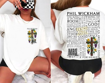 May include: White t-shirt featuring the text "PHIL WICKHAM" and a cross design with flowers and grapes. The back of the shirt has additional text and a similar cross design. The shirt is a casual wear item.