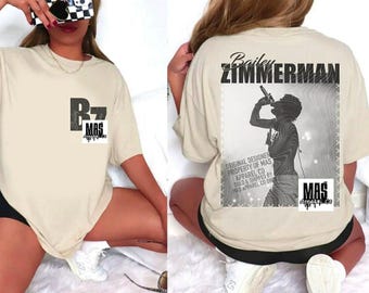May include: Beige short-sleeved t-shirt with a black and white graphic on the front and back. The front graphic features the letters "BZ" above "MAS". The back graphic features a singer and the name "Bailey Zimmerman".
