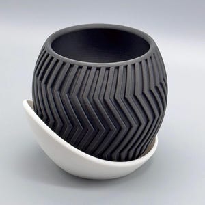 May include: A black ceramic planter with a geometric pattern and a white saucer. The planter is round and has a wide opening.