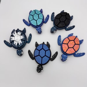 May include: Five colorful turtle figurines with textured shells and flippers are arranged on a white surface. The turtles feature various shell designs in shades of blue, black, white, and orange. Each turtle has a unique pattern.