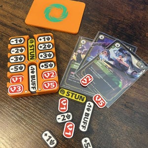 May include: A tabletop game accessory set featuring an orange plastic token organizer, a matching orange rectangular card, and several game cards. The organizer holds various white tokens with black and red text, including numbers and the words STUN and BUFF.