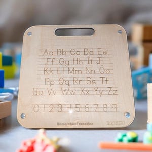 以下が含まれることがあります： 木製の教育用ボード。大文字と小文字の文字、数字、罫線が彫られています。ボードの上部にはハンドルがあり、銀色のネジで固定されています。「Homeschool creation」の文字が下部にあります。