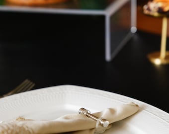 Op de afbeelding: Een wit keramiek bord met een beige linnen servet gevouwen en vastgezet met een zilveren servetring. Het servet heeft een delicate kantversiering.