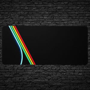 May include: Black rectangular desk mat with a colourful design. The design features a curved light blue line and three parallel diagonal lines in red, yellow, and green. The mat has rounded corners.