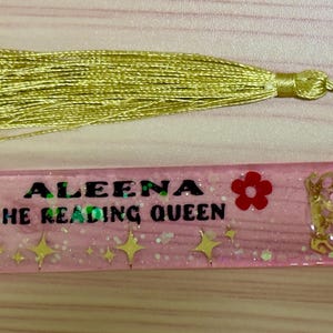 May include: A pink resin bookmark with a gold tassel. The bookmark features the text "ALEENA THE READING QUEEN", a unicorn graphic, red flowers, and gold stars. The bookmark is approximately 6 inches long.