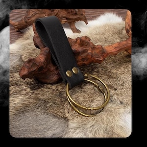 Peut inclure: Une boucle de ceinture en cuir noir avec deux rivets en laiton et un double anneau attach&eacute;. La boucle est sur une surface de fourrure brune et blanche.