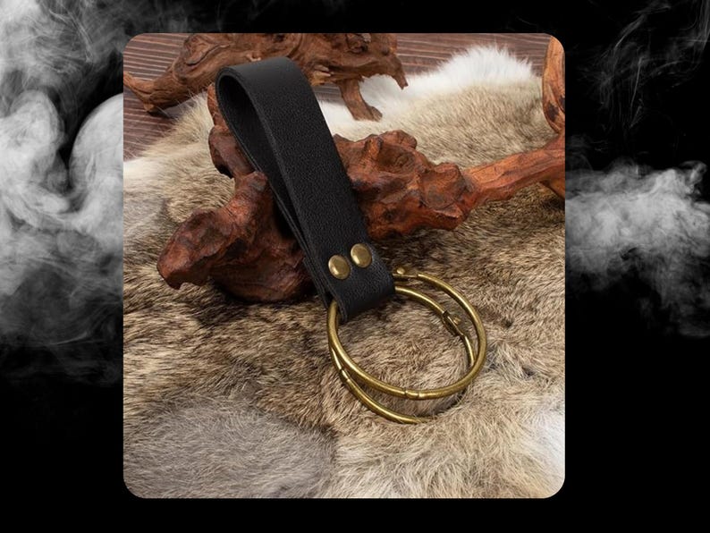 Peut inclure: Une boucle de ceinture en cuir noir avec deux rivets en laiton et un double anneau attach&eacute;. La boucle est sur une surface de fourrure brune et blanche.