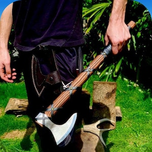 May include: A brown-handled axe with a black leather wrap and metal accents. The axe is held by a person wearing black pants and a black shirt.