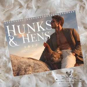 May include: A 2027 wall calendar titled Hunks & Hens featuring a photograph of a person wearing a blazer and light-colored pants, sitting with a brown chicken. The cover includes the text Hunks & Hens 2027 and Living on Madison.