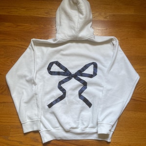Könnte beinhalten: Weißer Kapuzenpullover mit einem marineblauen Schleifendesign auf dem Rücken. Die Schleife besteht aus einem gemusterten Stoff. Der Pullover hat eine Kapuze mit Kordelzug und gerippte Bündchen und Saum. Ein lässiges, bequemes Kleidungsstück.
